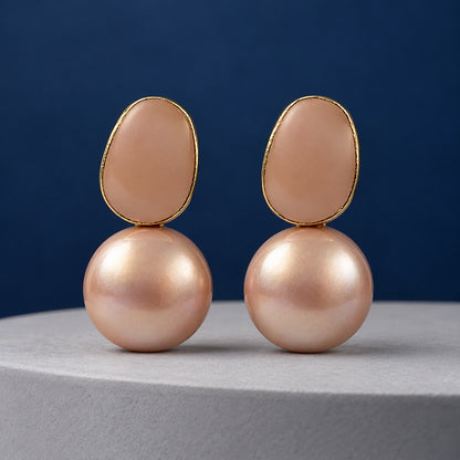 Blush Pebble Pearl Drops gold-tone earrings with blush pebble top and rose-pink faux pearl on a cream pedestal